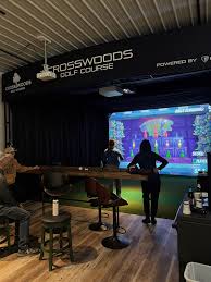 A golfer's perspective of a professional indoor golf simulator bay with a large projection screen displaying a scenic golf hole, professional turf hitting mat, and adjacent seating area.